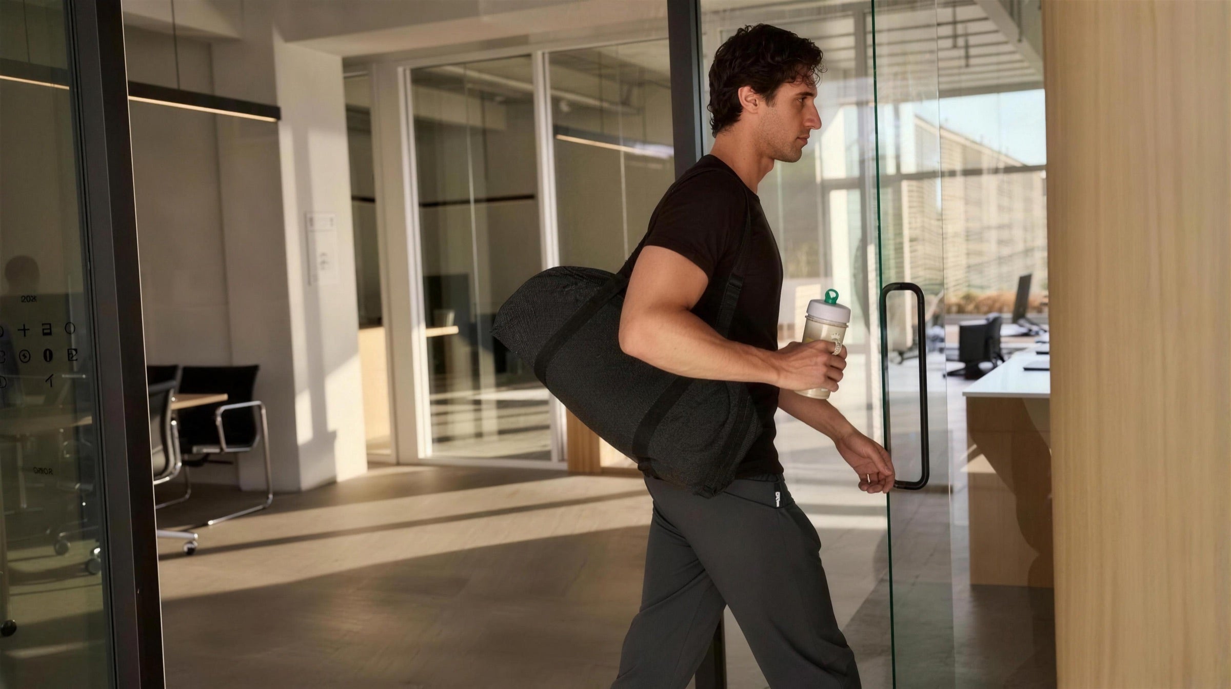 A man arriving at work in the morning, gym bag over his shoulder and a shaker in his hand.
